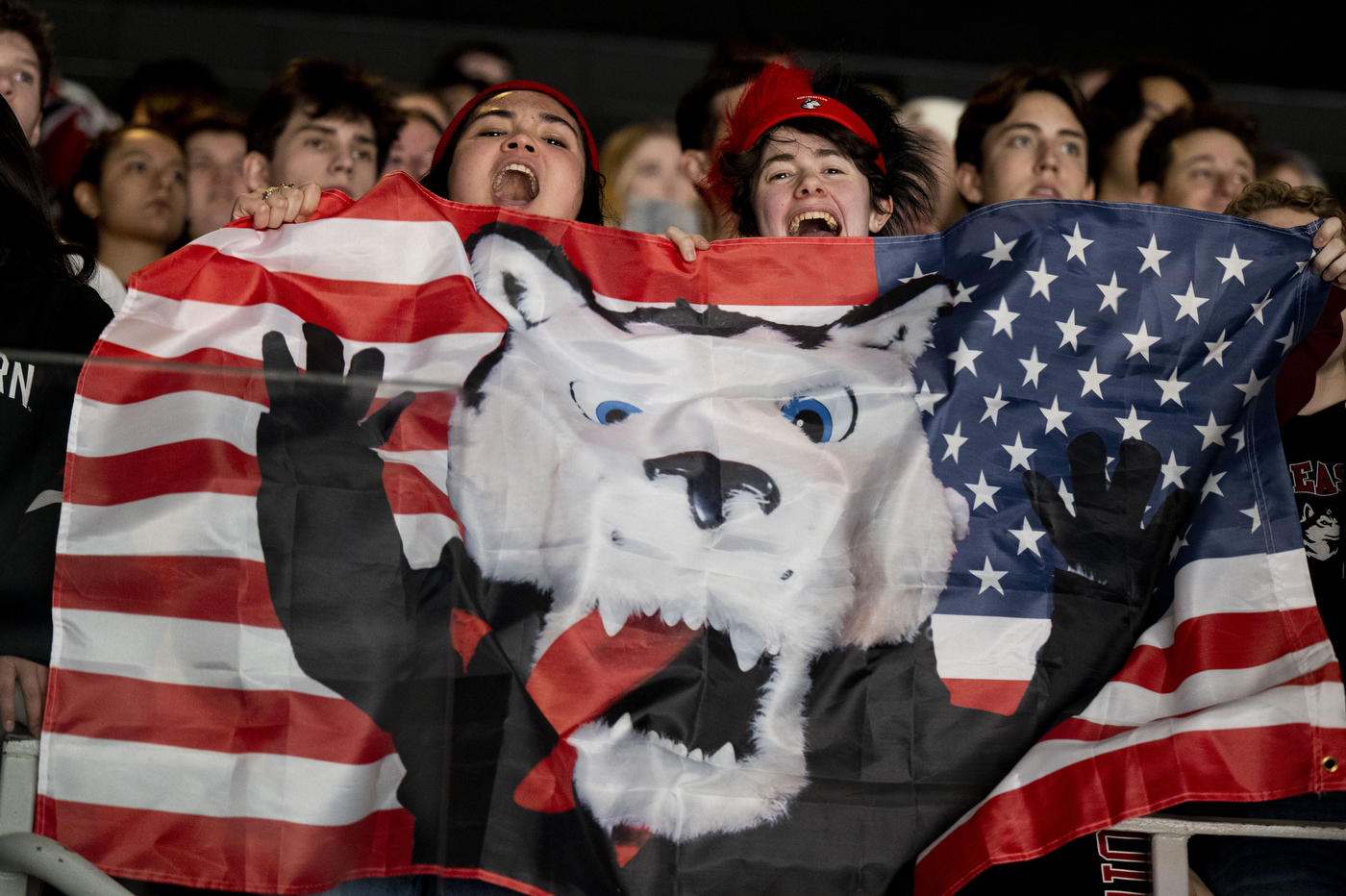 Cheering Northeastern fans hold a large American flag banner featuring a husky graphic during the Men’s Beanpot semifinal at TD Garden.