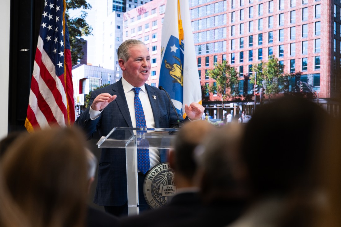 A person adressing the crowd stands a podium, with two flags behind him.