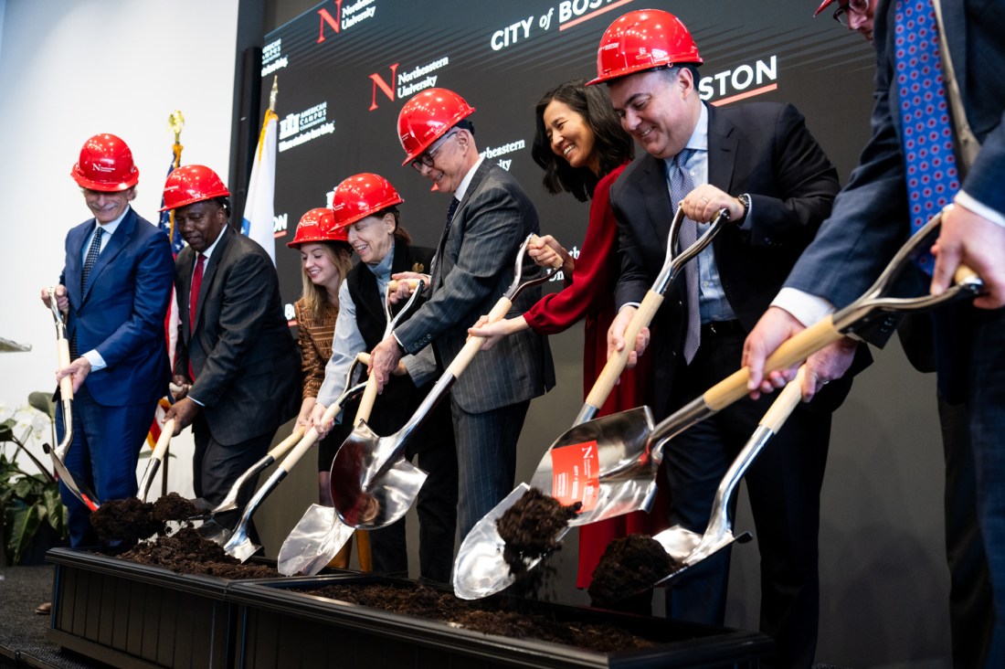 Several people wearing hard hats use shovels to toss dirt into a container.