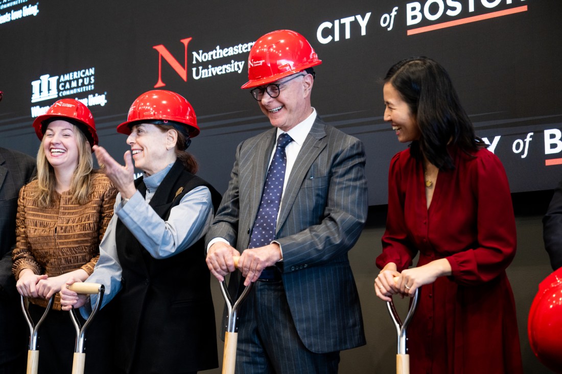 The university president wears a red hard hat, standing on stage with the mayor of Boston and others, while holding shovels.