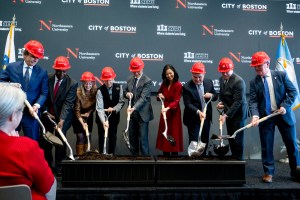 A group of people including Mayor Michelle Wu and President Aoun wearing red hard hats take shovels to break ground on the new residence hall