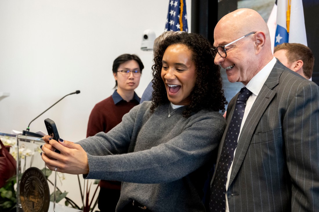A side profile shows the university president on stage in front of a microphone, with another person.