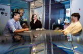 Brooke Williams, librarian, and students Quoc Hung Le and Alexander Walulik talk together in a modern-looking glass office with some colorful posters and stickers.