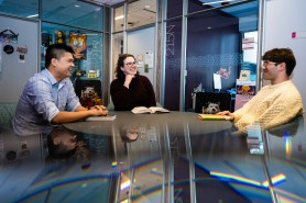 Brooke Williams, librarian, and students Quoc Hung Le and Alexander Walulik talk together in a modern-looking glass office with some colorful posters and stickers.