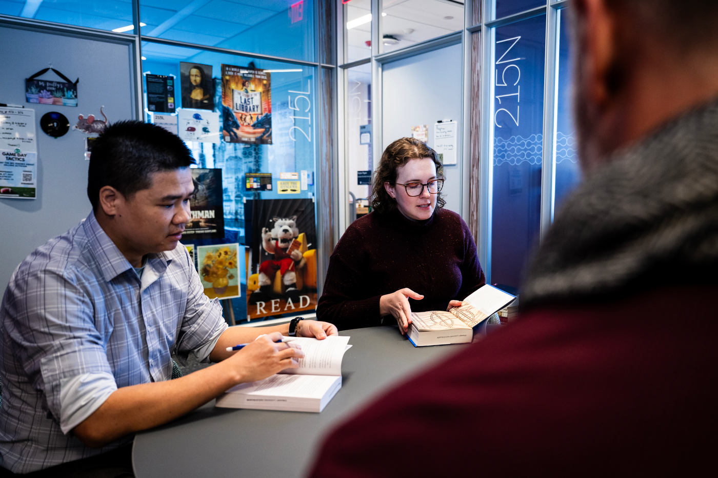 Brooke Williams and Quoc Hung Le reading a book together in a modern-looking glass office with stickers and posters in the background.