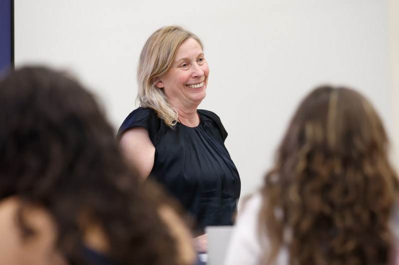 Juliana Saphr, wearing a black sleeveless top, smiles while teaching and engaging with students who are visible from behind in the foreground, in a bright classroom setting with a presentation screen partially visible on the left.