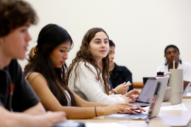 Students sit at classroom tables engaged in discussion, with focus on a student in a white top looking thoughtfully forward while classmates listen and participate in conversation in a bright, minimalist classroom setting.