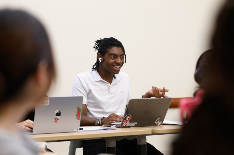 A smiling student in a white collared shirt actively participates in a classroom discussion, gesturing with hands while speaking to classmates visible from behind in the foreground, in a bright minimalist classroom setting.