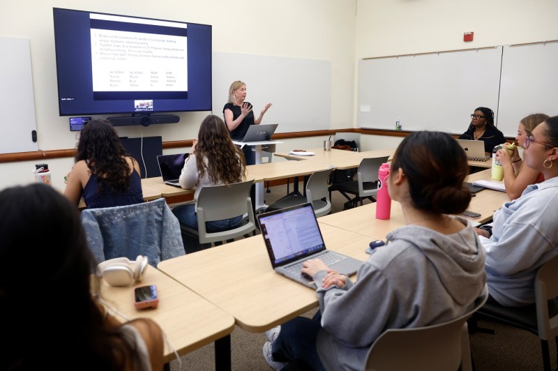 Juliana Saphr stands at the front of a classroom gesturing while teaching, with a large presentation screen displaying text behind her, as students sit around curved tables facing her with laptops, water bottles, and notebooks, in a bright modern classroom with whiteboards visible on the walls.