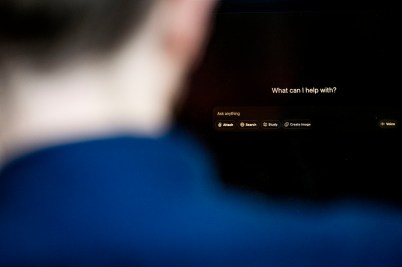Looking over the shoulder of someone wearing a blue shirt, a computer screen reads "What can I help with?"