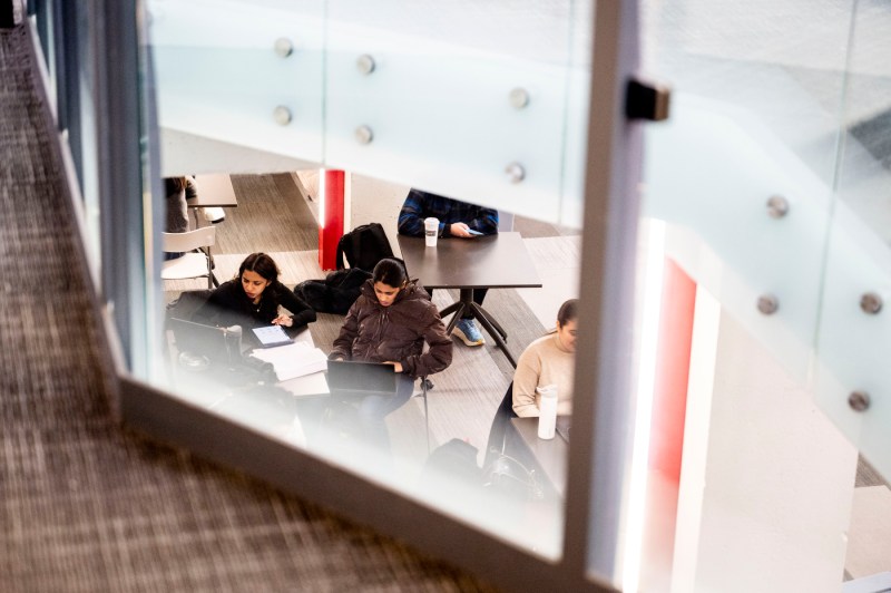 Shot through glass, students sit at square tables while working on laptops and using their phones. 
