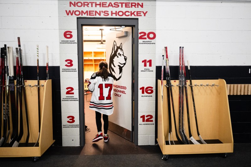 Ella Tapp, who is wearing a Northeastern jersey with the number 17 and her last name, walks through the door for the Northeastern women's locker room. Above the door, a sign reads, "Northeastern women's hockey."