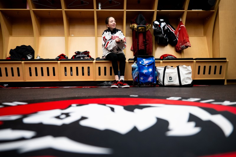 Ella Tapp, wearing a Northeastern hockey jersey, sits on a bench in the Northeastern women's locker room.