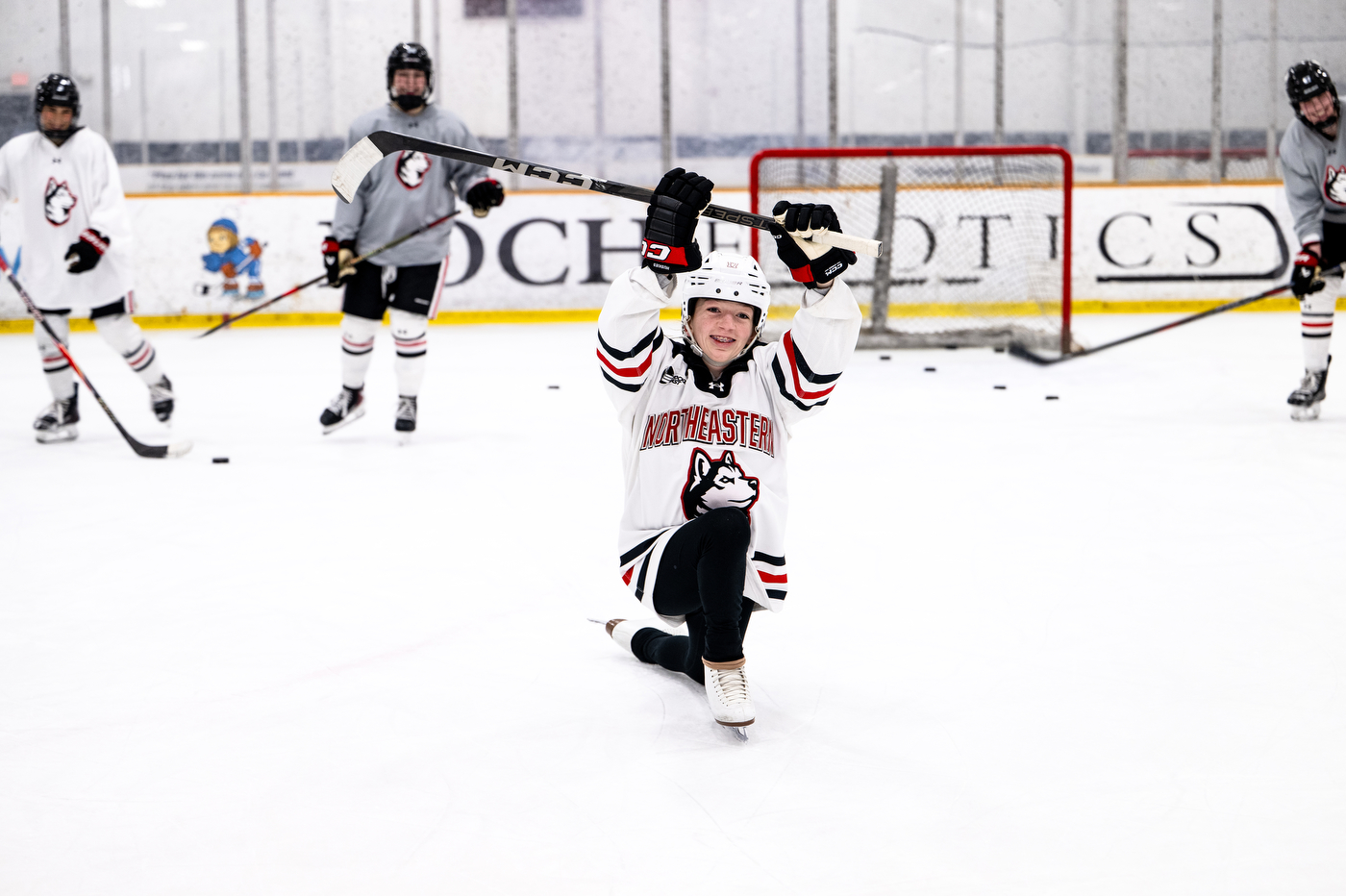 Ella Tapp, wearing Northeastern hockey gear, kneels down on the ice and holds up her hockey stick in celebration. 