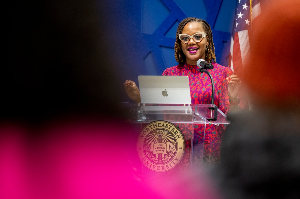 A woman in glasses and bright pink dress speaks into a microphone at a podium. There is an Apple laptop in front of her and an American flag behind her.