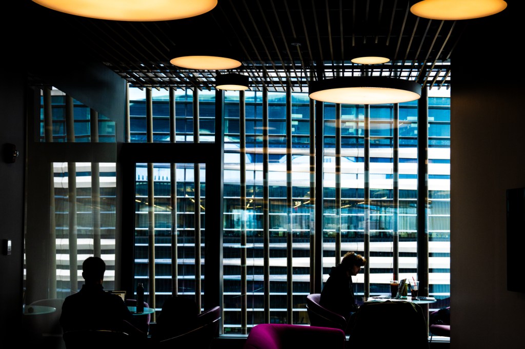 Silhouettes of students study at tables in a dramatic modern library space featuring floor-to-ceiling illuminated bookshelves arranged in a grid pattern, with warm overhead lighting and blue-tinted windows visible in the background.