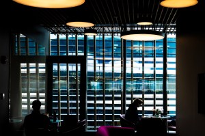 Silhouettes of students study at tables in a dramatic modern library space featuring floor-to-ceiling illuminated bookshelves arranged in a grid pattern, with warm overhead lighting and blue-tinted windows visible in the background.
