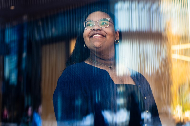 Portrait of Amelia Brooks wearing a blue cardigan over a black shirt and glasses. Amelia is looking off into the distance beyond the camera.
