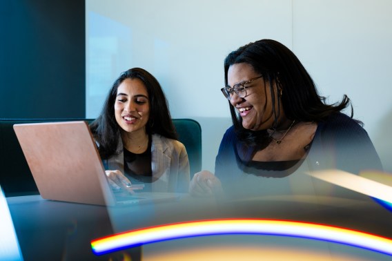 Arshia Mathur and Amelia Brooks sitting side by side at a table in front of one laptop. They are both looking at the laptop and smiling or laughing