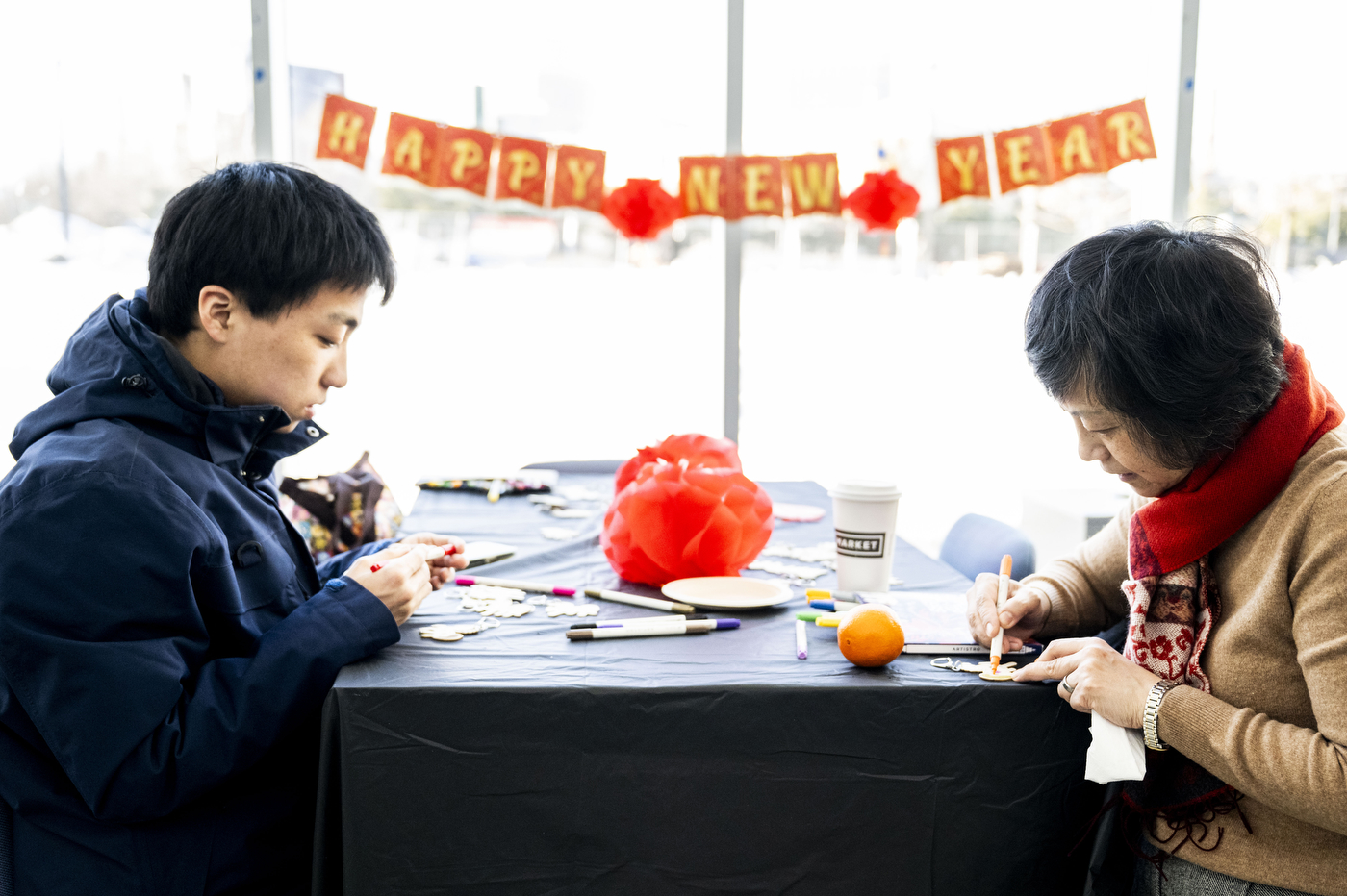 As part of a Lunar New Year celebration, two people work on crafts at a table. A banner that reads, "Happy New Year" hangs from a window in the background. 