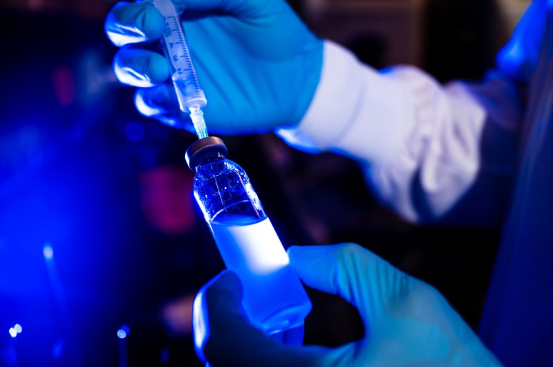 Gloved hands handling small glass bottles containing liquid samples in a laboratory setting.