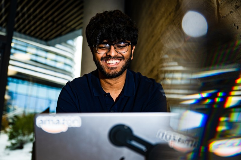 A student wearing glasses smiles while looking down at a laptop screen.