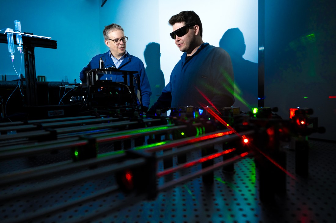 Someone in glasses and a blue labcoat stands next to another person in sunglasses. In front of them is a table with green and red lasers.