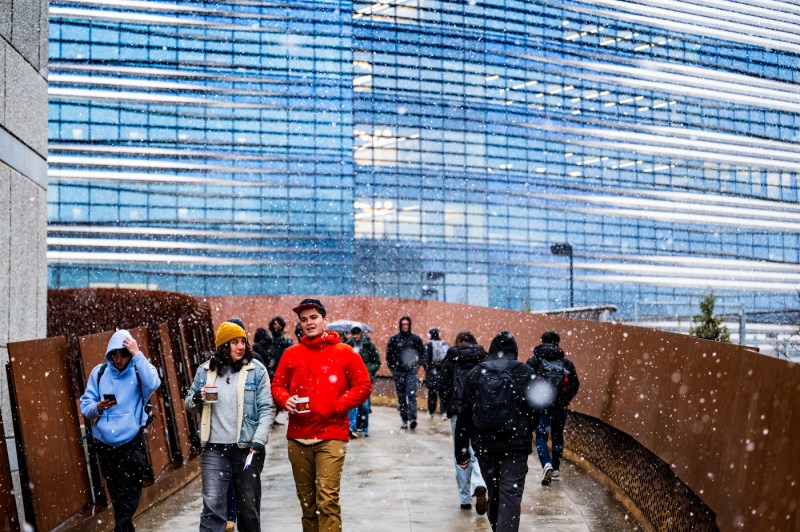 Students walk outside on a bridge between buildings as snow falls.