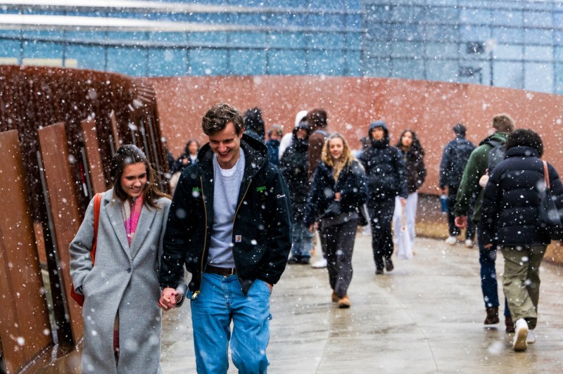 A couple holding hands, and other people, walk across a bridge between buildings as snow falls.