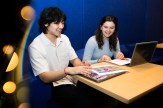 Dylan Kao and Lucy Paolini sit at a table reviewing colorful printed materials, with a laptop open, against a blue background with warm bokeh lighting on the left.