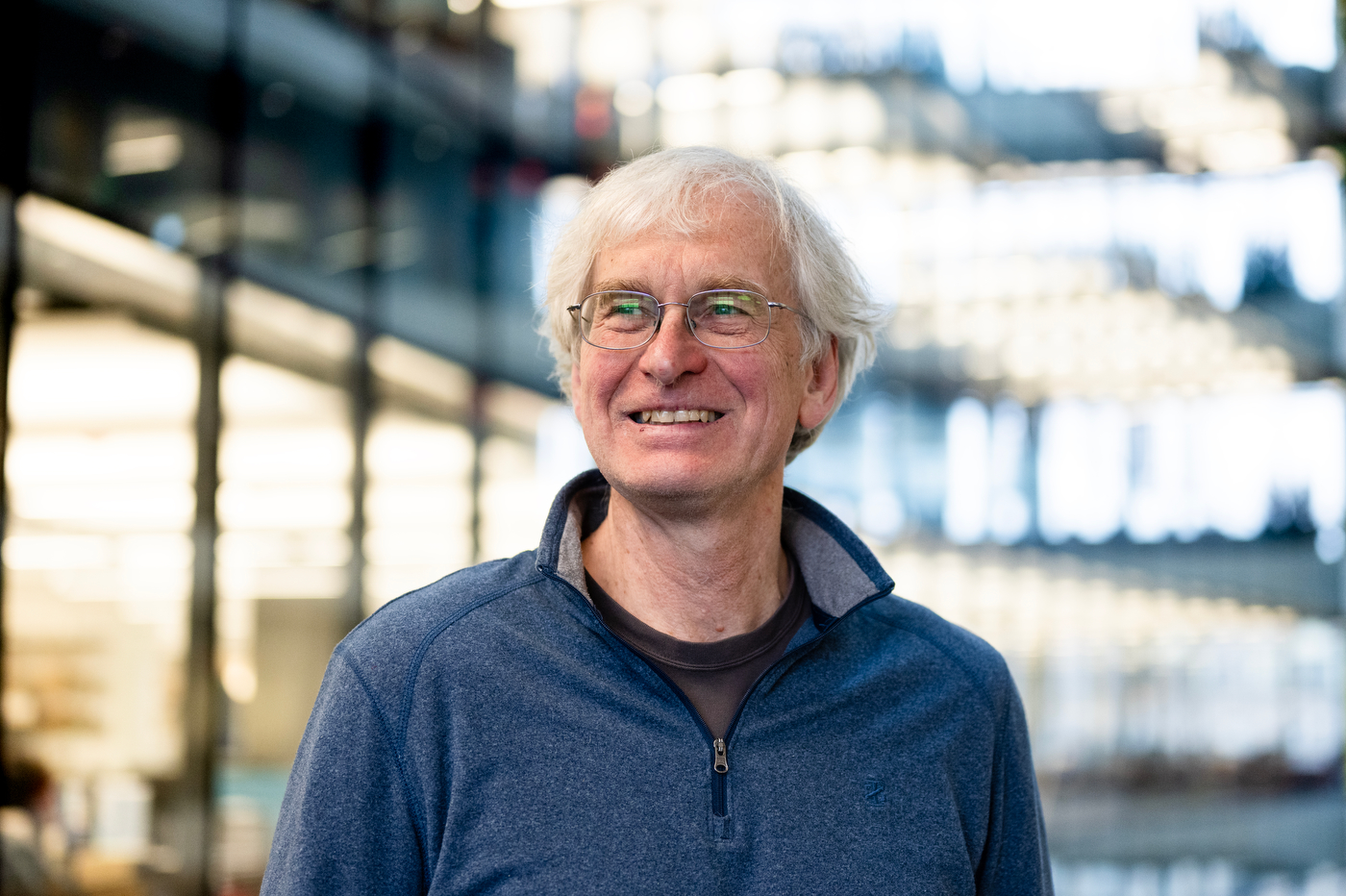 A man with grey hair and glasses smiles posing for a portrait inside an academic building with rows of glass windows behind him.
