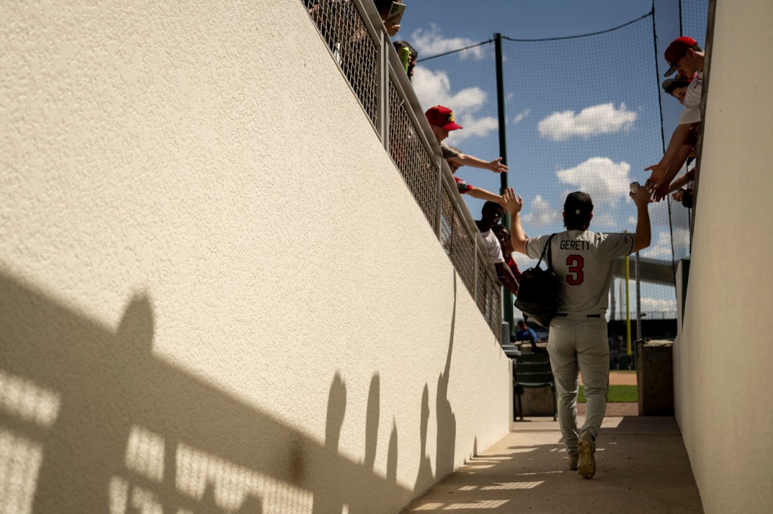 A baseball player in a gray uniform walks down a narrow concrete corridor toward the field, raising his arm to greet fans leaning over a railing above him.