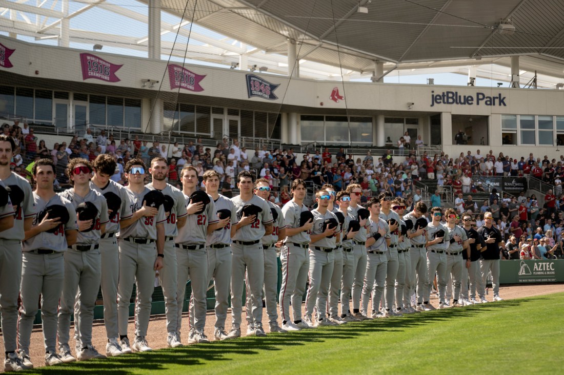 Northeastern's baseball team in matching gray uniforms stands shoulder to shoulder with their hats over their hearts along the outfield grass during a pregame ceremony, with a packed stadium behind them at jetBlue Park.
