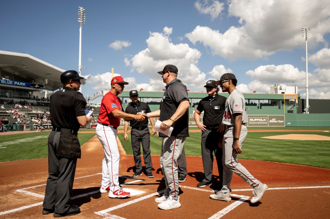Players, coaches, and umpires gather at home plate exchanging handshakes before a game, with the infield dirt, green outfield wall, and bright blue sky in the background.