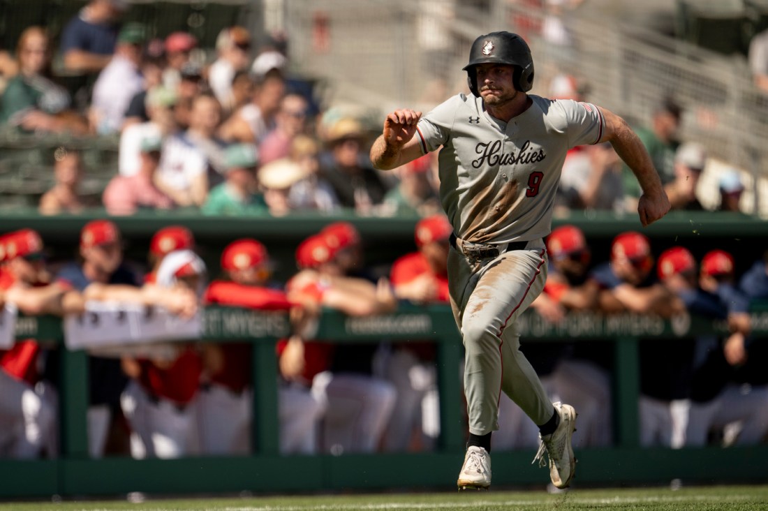 A baseball player in a gray uniform runs the bases after making contact, while players in the opposing team’s dugout watch from the background.