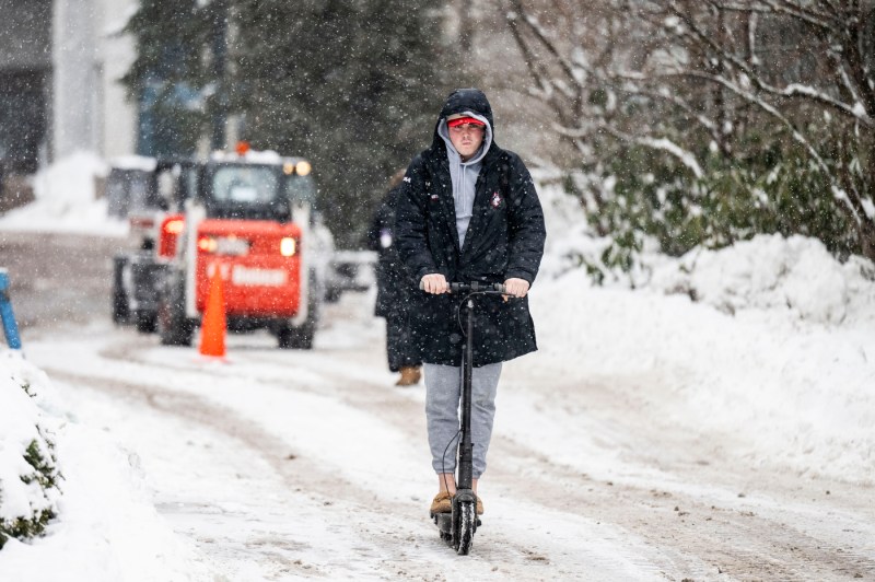 A person dressed in heavy winter clothing rides an electric scooter through streets filled with snow.