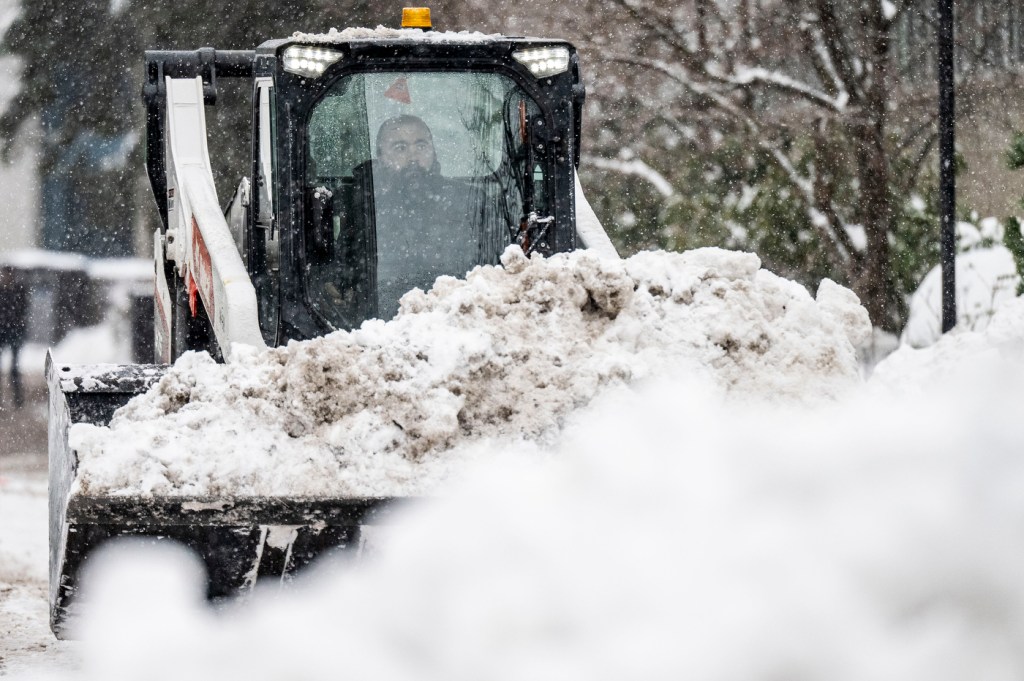 A Bobcat, driven by a man, hauls a bucket full of snow on a street, surrounded by mountains of snow.