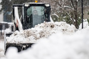 A Bobcat, driven by a man, hauls a bucket full of snow on a street, surrounded by mountains of snow.