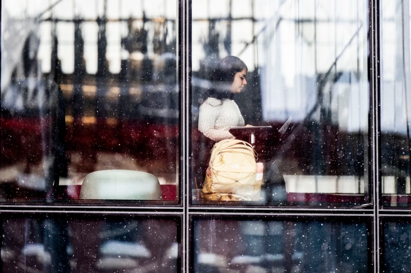 Through a window, a person uses their laptop while sitting at a table.