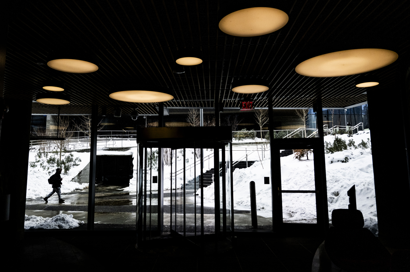 Inside a darkened exterior as the camera faces brightly lit windows. Outside, a person walks towards a staircase, surrounded by snow banks.