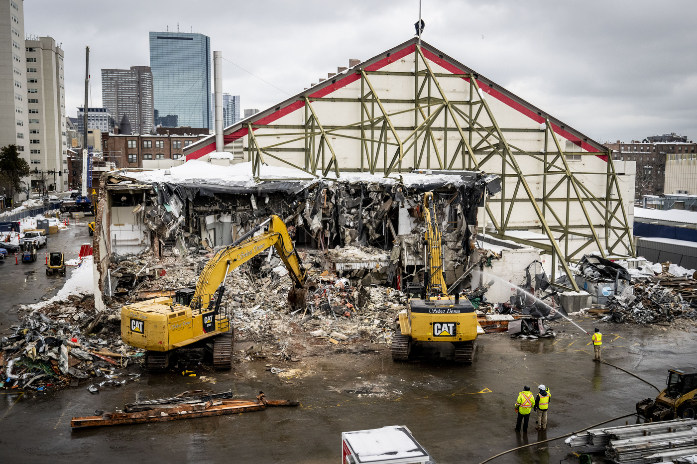 An active construction site where construction workers wearing bright neon safety vests coordinate alongside heavy machinery to bring down a building, with debris and rubble visible throughout the surrounding area.