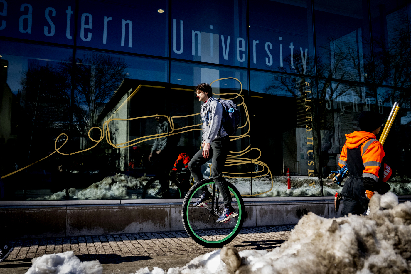 A student pedals a unicycle on a sidewalk in front snow piles and a Northeastern University building