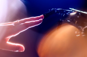Close-up of a human hand reaching out to touch a robotic hand, illuminated by warm, glowing light against a dark background, evoking a moment of connection between human and technology, such as a human using an LLM chatbot.