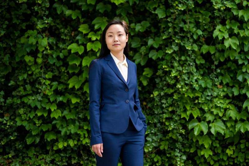 Amy Pei wearing a blue suit and white shirt standing in front of a wall of green foliage.