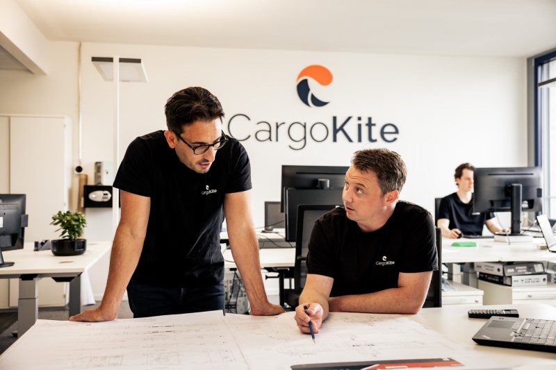 Employees wearing black t-shirts work together around a desk inside a CargoKite office, reviewing large paper plans on a tabletop. 