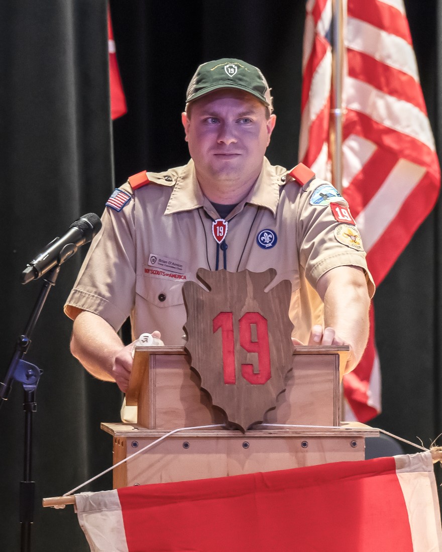 A man in khaki Scouting uniform, hat and regalia stands behind a wooden podium with a microphone on a stand to his right and American flag to his left.