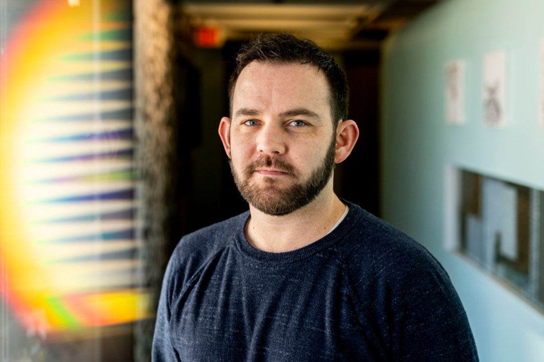 Portrait of Brennan Klein wearing a dark long sleeve shirt standing in a hallway with turquoise walls. 