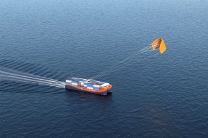 A cargo ship moving through open water as a large orange kite pulls it forward using wind power.