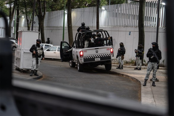 Armed personnel in tactical gear surround a police pickup truck on a tree-lined street outside a white fence, photographed from inside a vehicle with the window frame visible in the foreground.