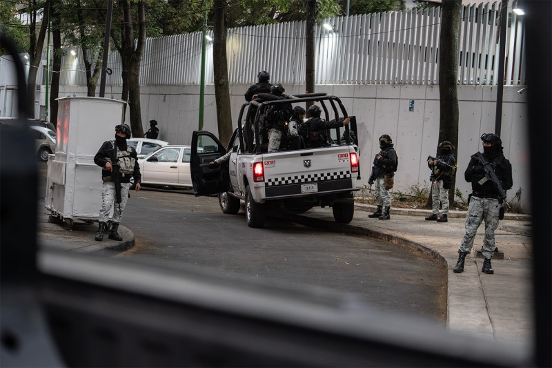 Armed personnel in tactical gear surround a police pickup truck on a tree-lined street outside a white fence, photographed from inside a vehicle with the window frame visible in the foreground.